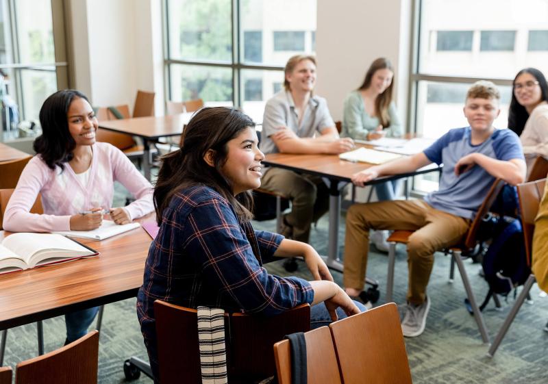 A group of students sits around tables in a bright classroom, engaged in discussion and note-taking, with books scattered on the desks.